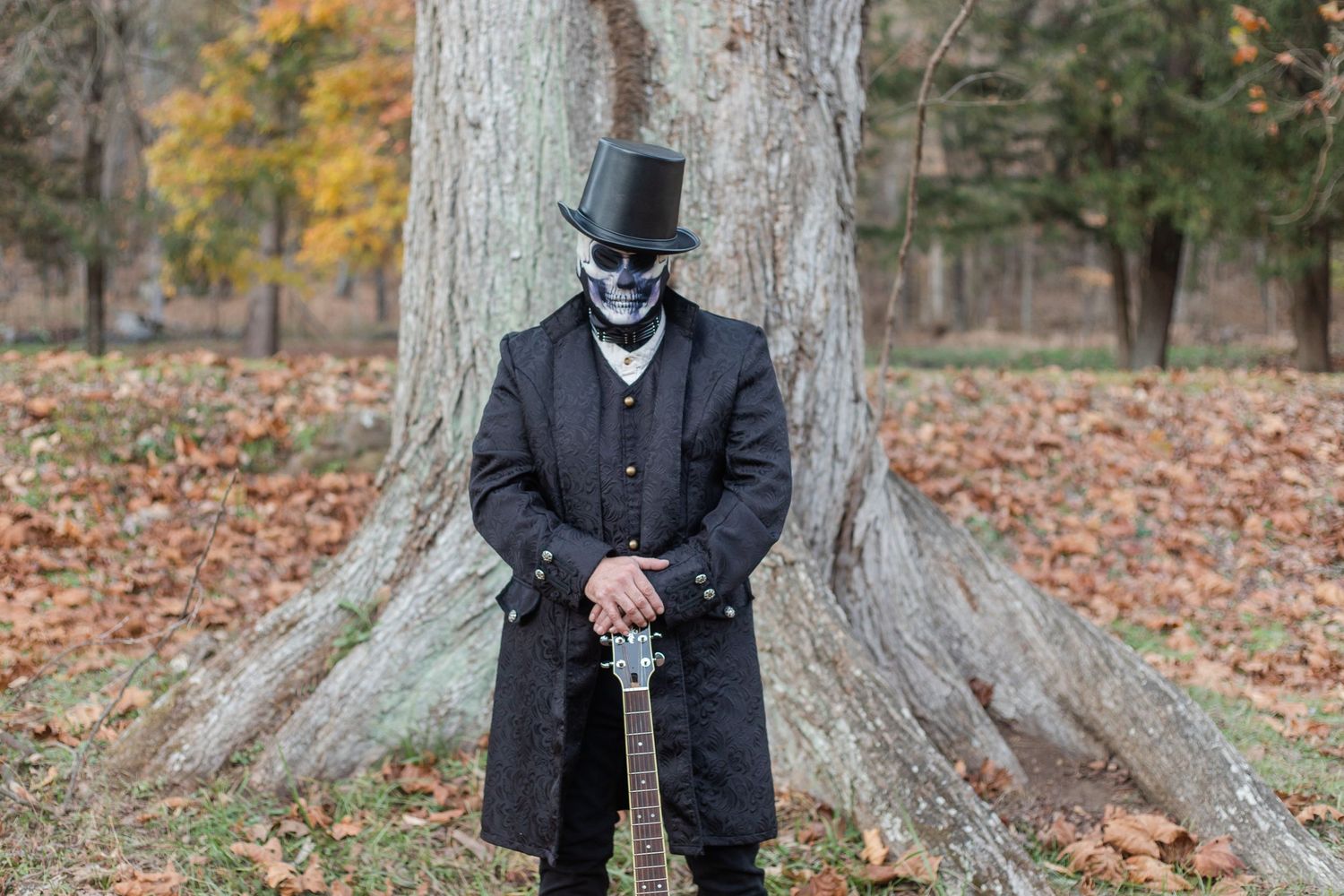 A person in formal attire poses against a large tree trunk in autumn.