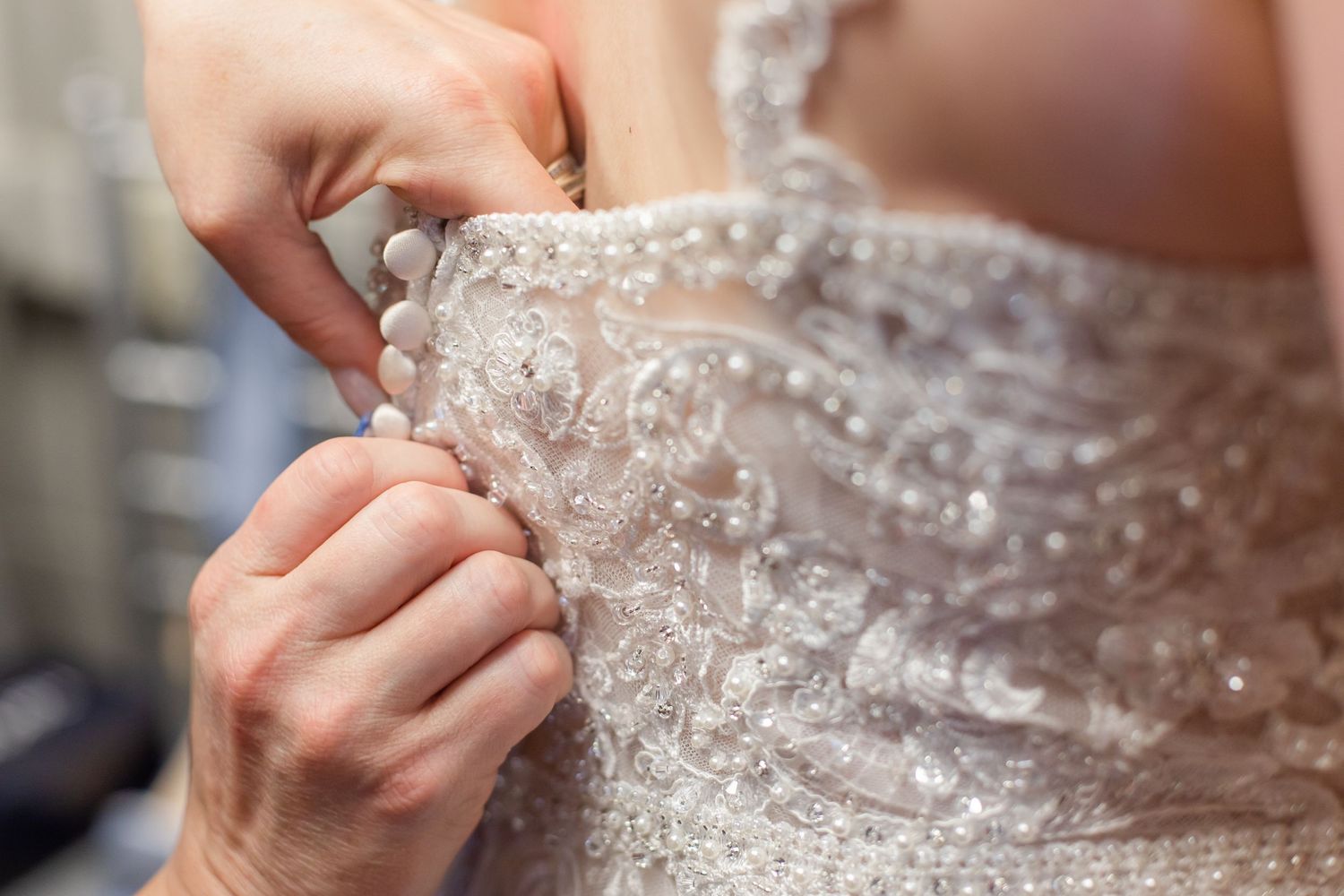 Close-up detail of hands fastening delicate buttons on a beaded lace wedding dress.