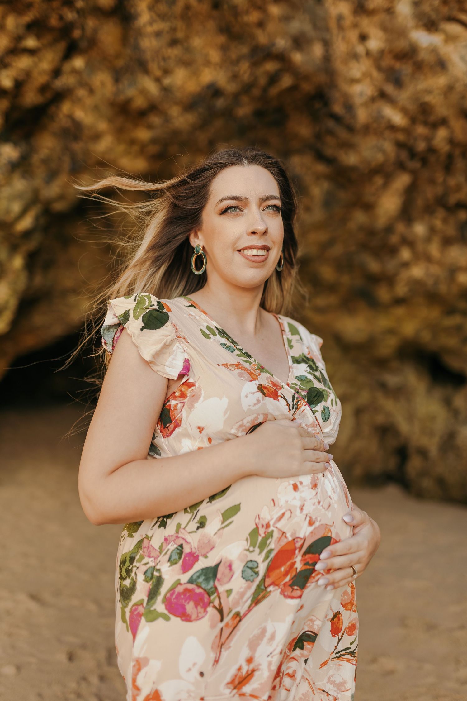 Pregnant woman in floral dress cradles baby bump on sandy beach with rocky backdrop.