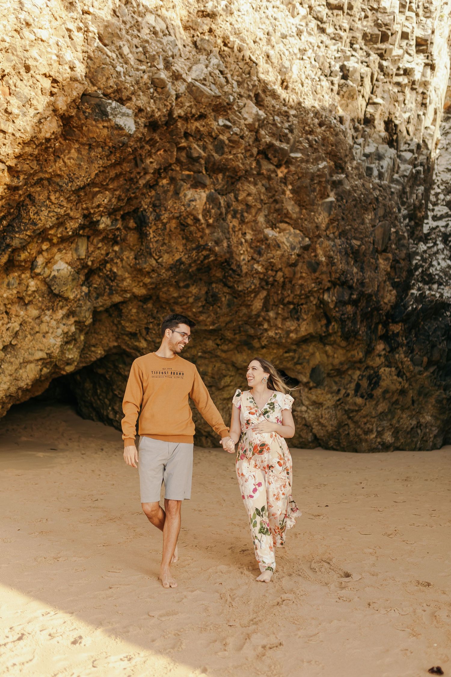 Couple holding hands walking barefoot on sandy beach near rocky cave wall.
