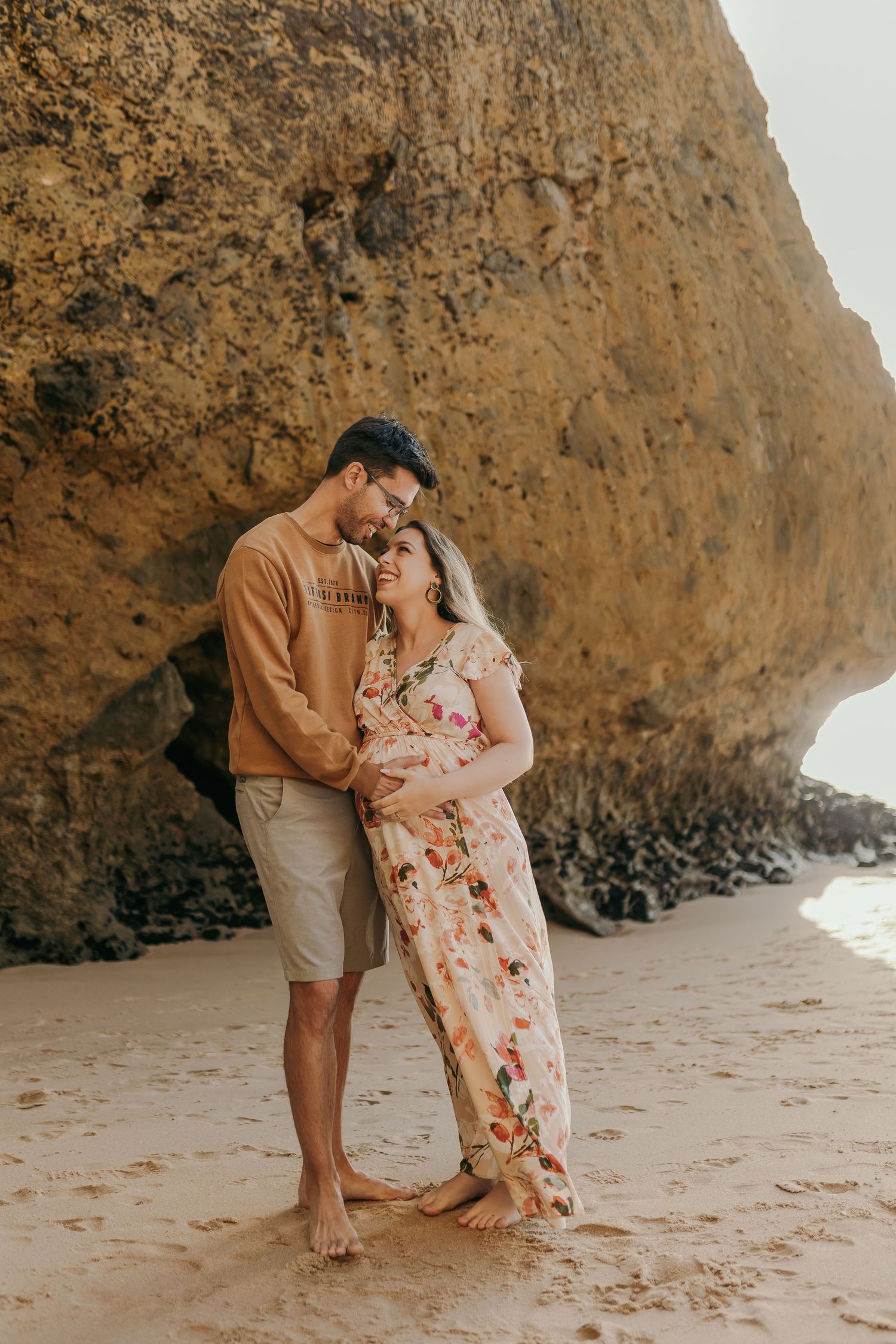 Pregnant couple embracing tenderly on beach with rocky cliff backdrop.