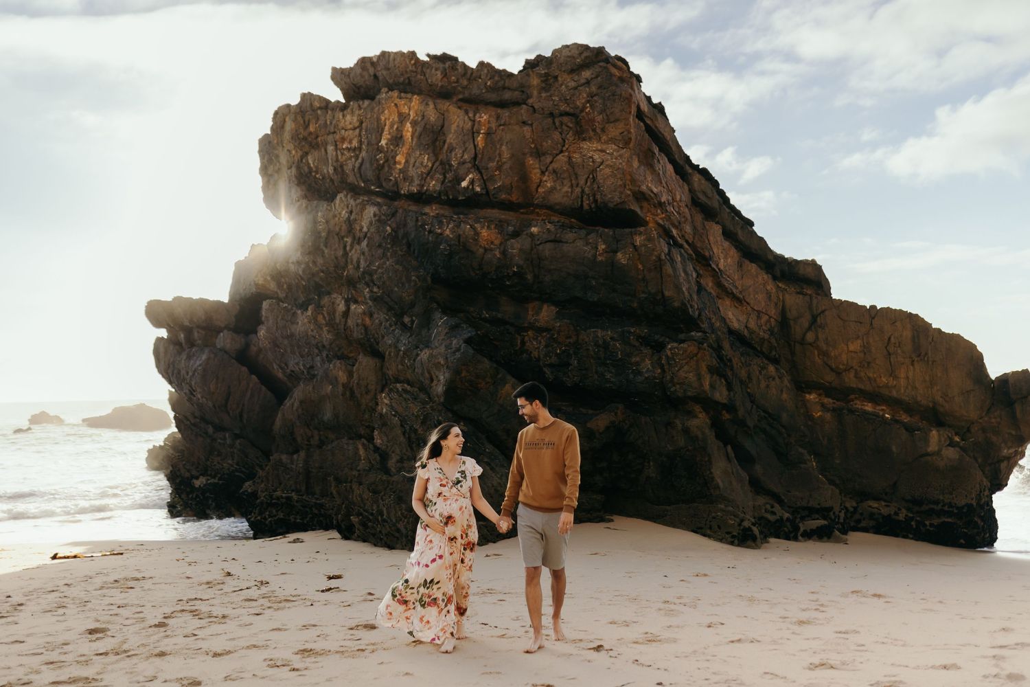 Couple walking hand in hand on beach with dramatic sunlit rock formation.