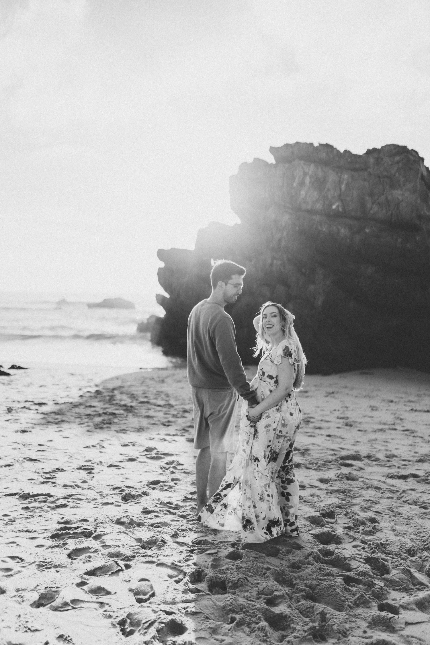 Black and white photo of couple holding hands on sandy beach by rocks.