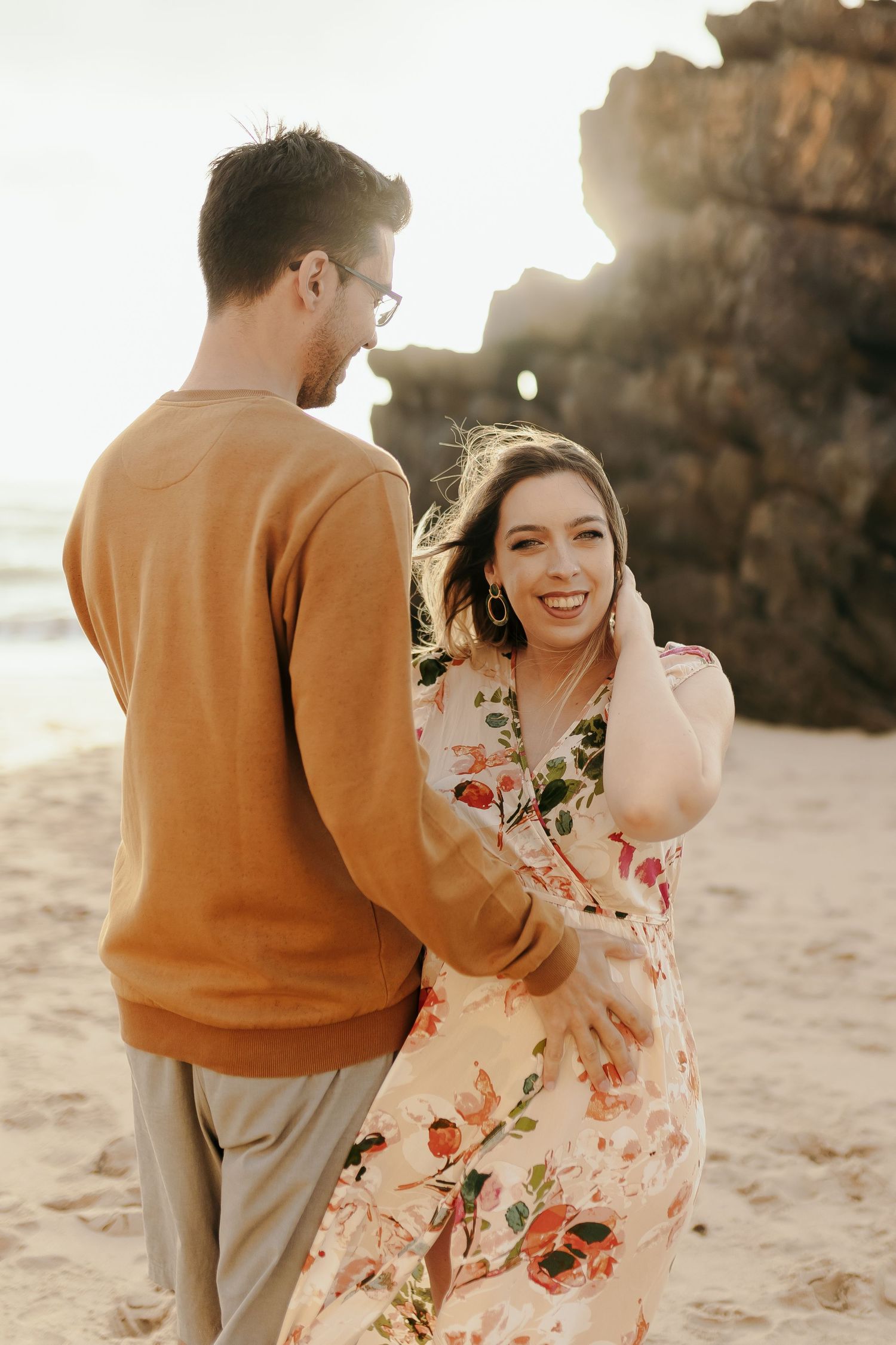 Smiling pregnant woman in floral dress embracing partner on sunny beach.