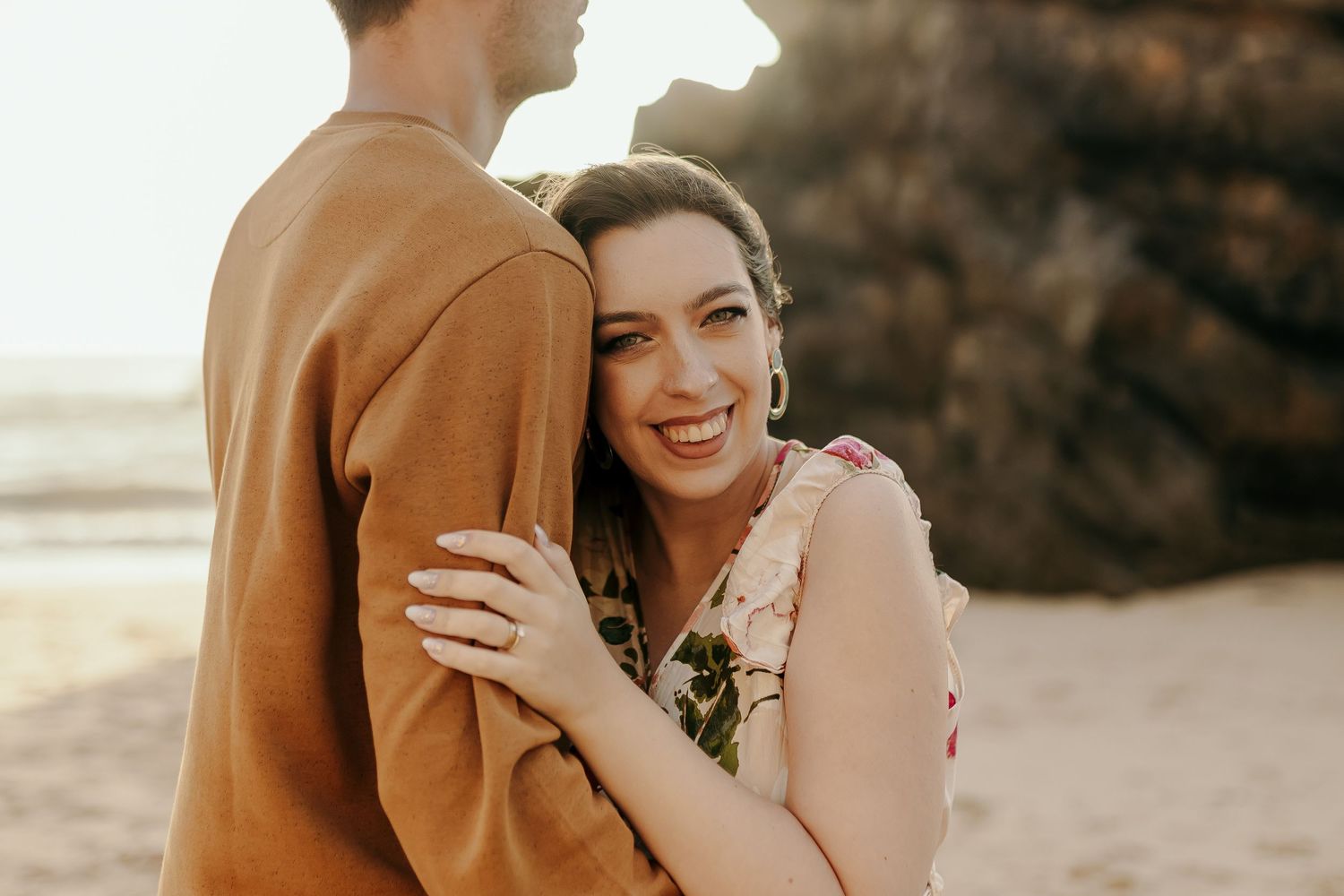 Happy woman in floral dress hugging partner on golden sunset beach.
