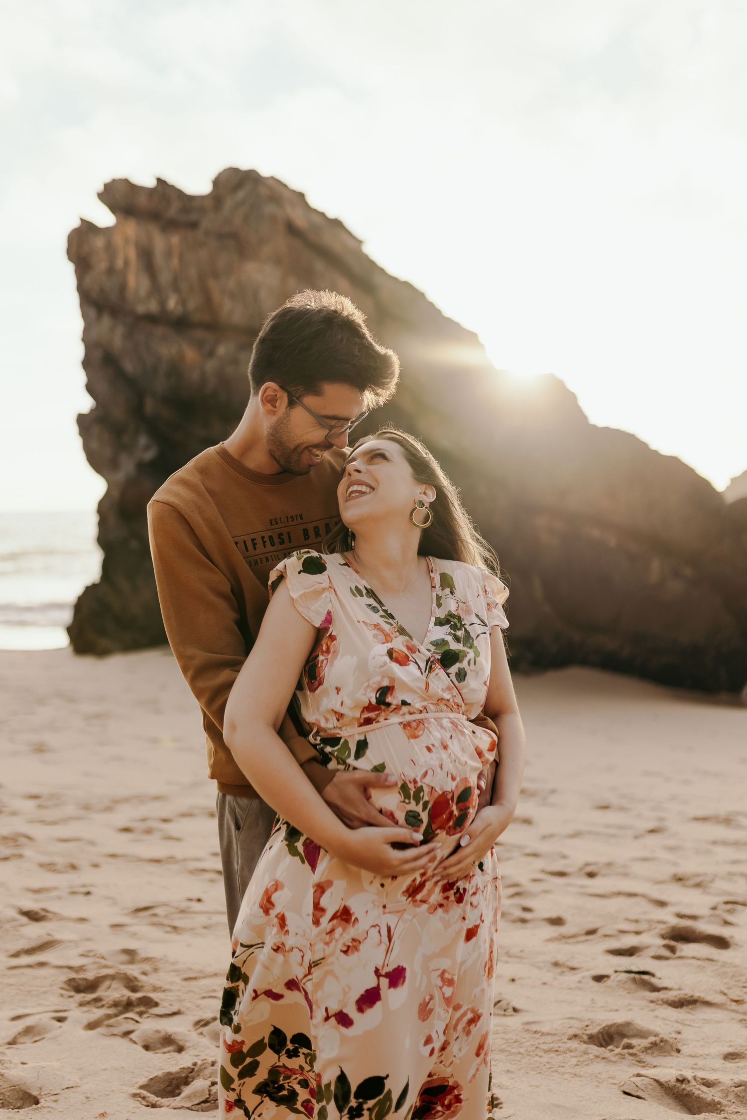 Man kissing pregnant woman's cheek while holding her belly on sunny beach.