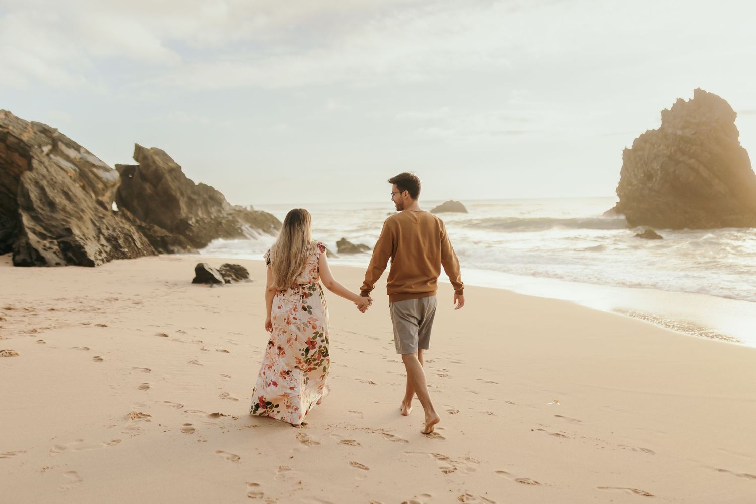 Couple walking hand in hand along ocean shoreline with rocky cliffs.