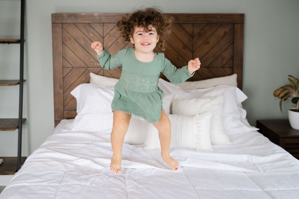 Child in green shirt jumping playfully on white bed with wooden headboard.