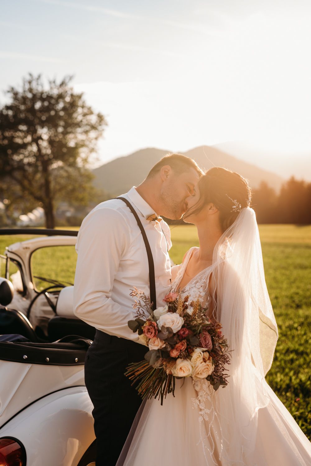 Romantic sunset wedding portrait with couple by vintage car in countryside setting.