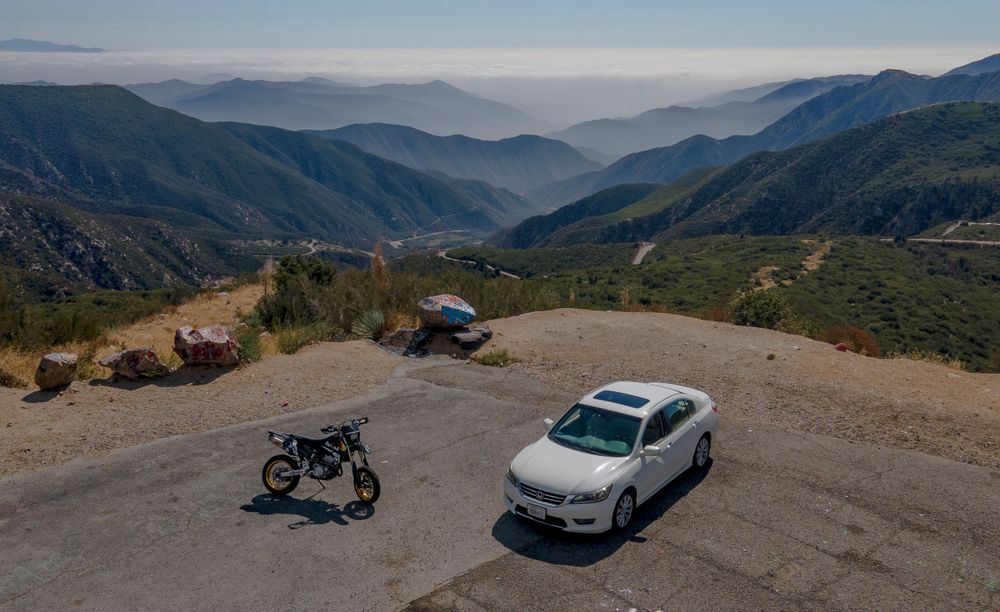 Aerial view of a motorcycle and white sedan parked on a mountain overlook with scenic valley views.