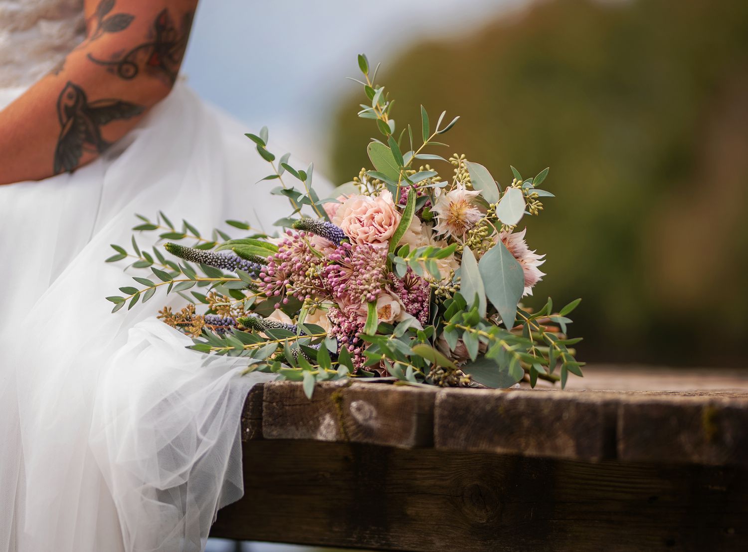 Bride holding rustic wildflower bouquet with eucalyptus and pink blooms, tattoo visible on arm.