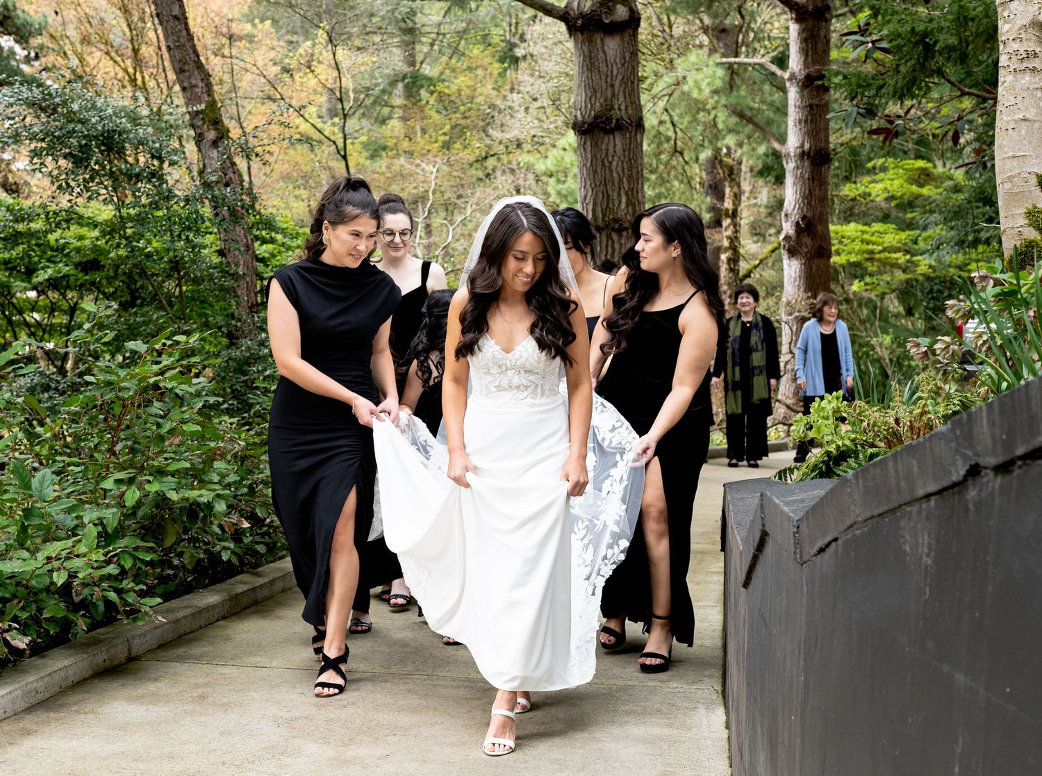 Bridesmaids in black dresses helping the bride with her white gown in a garden pathway.