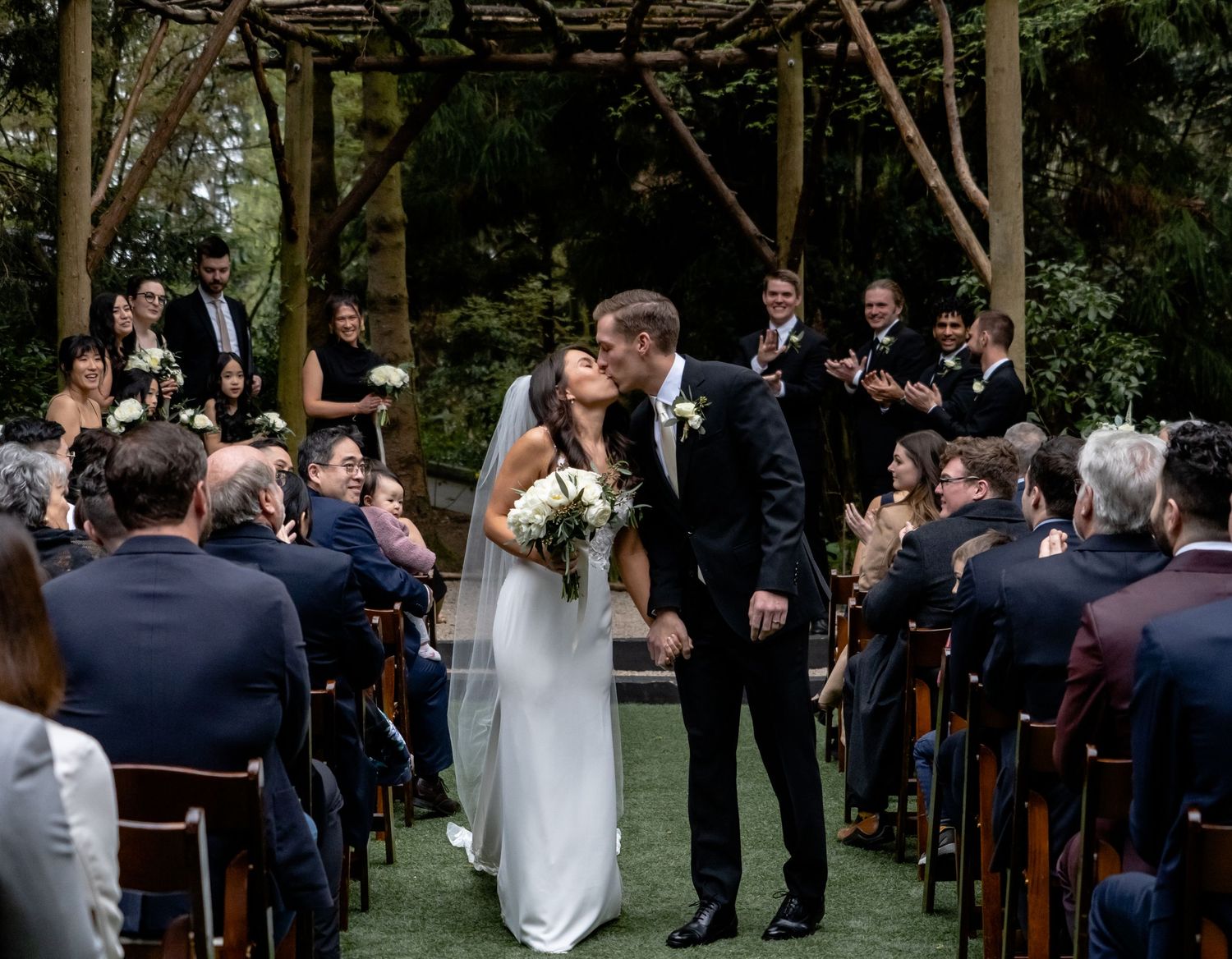 Outdoor garden wedding ceremony under wooden arbor with seated guests watching couple exchange vows.