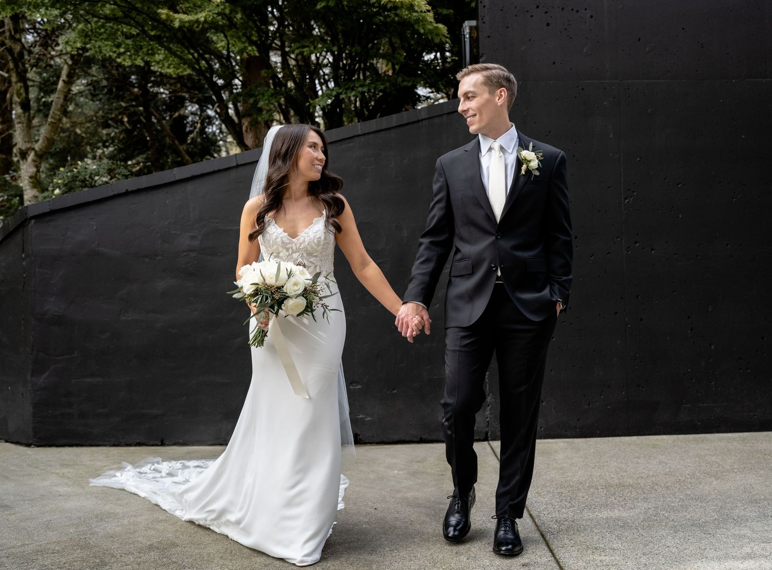 A couple in wedding attire walking and holding hands against a modern black wall backdrop.