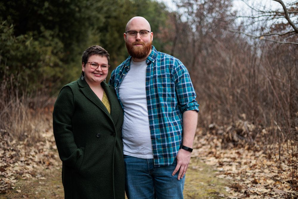 A couple poses together against a backdrop of trees and fallen leaves during an autumn photoshoot.