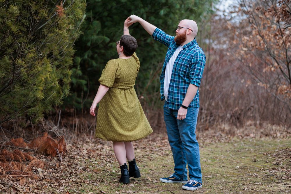 A couple embraces while standing in a wooded area with fallen leaves on the ground.