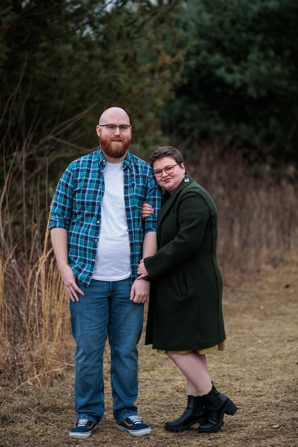 Two people pose together on a dirt path surrounded by autumn foliage and trees.