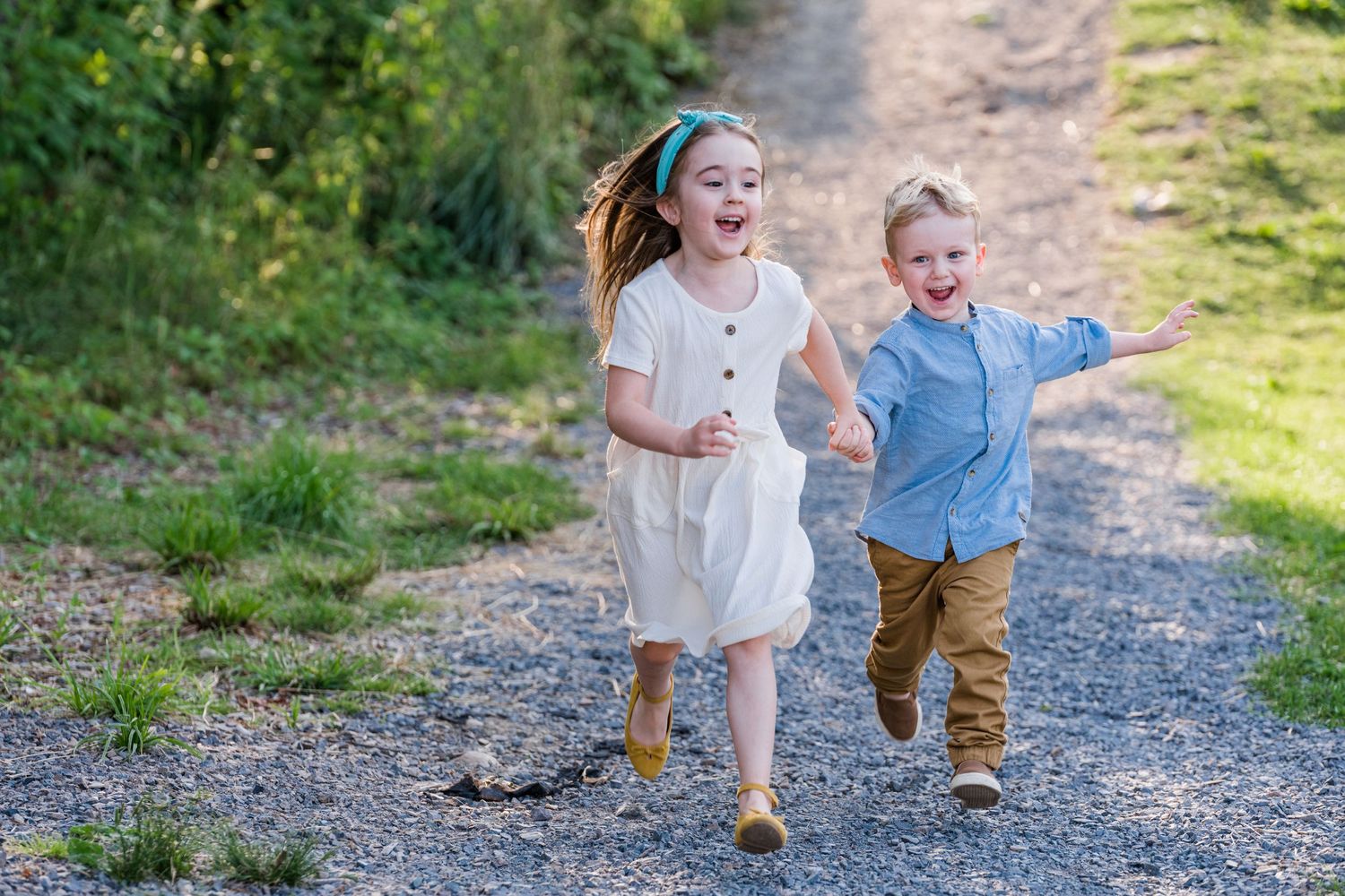 Two young children running playfully together on a gravel path surrounded by nature.