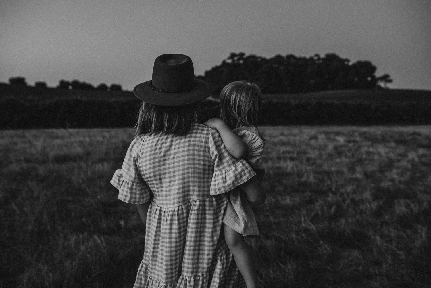 Black and white image of mother and child looking at sunset in field.