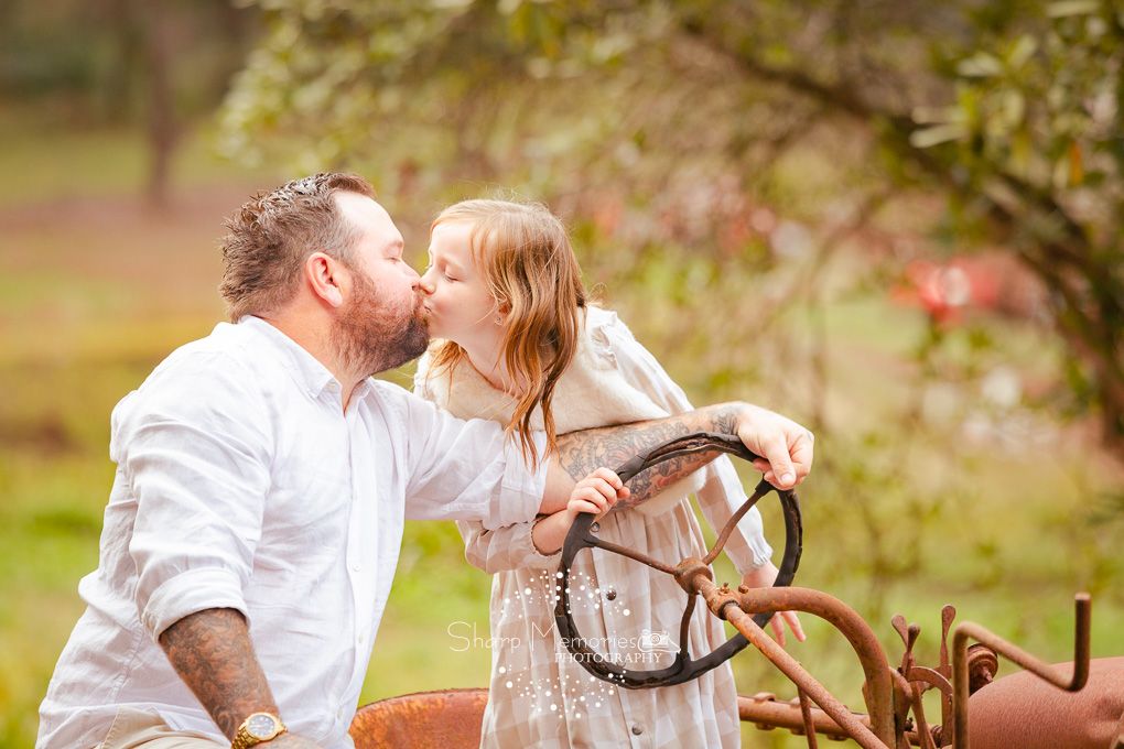 A daddy and daughter share a kiss by a rustic metal farm gate in a garden setting.