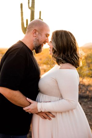 Maternity portrait of a pregnant mom and her husband touching noses in front of a cactus with a bright golden glow behind them in Phoenix.