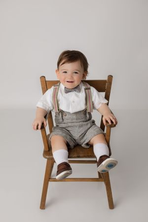One year portrait for this dapper little baby boy sitting on a wooden chair with a simple white backdrop for his first birthday photoshoot.