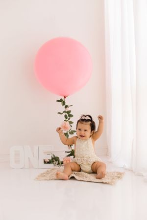 Happy baby girl smiles for her one year portrait session in the bright white studio with a large pink balloon in the studio.
