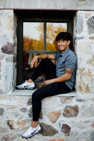 Senior graduate teenage boy sits in the window of an urban location and smiles at the camera for his senior portrait session.