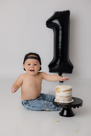 Handsome dude in jeans sits in front of a white backdrop with a white cake and black one balloon for a simple studio cake smash session.