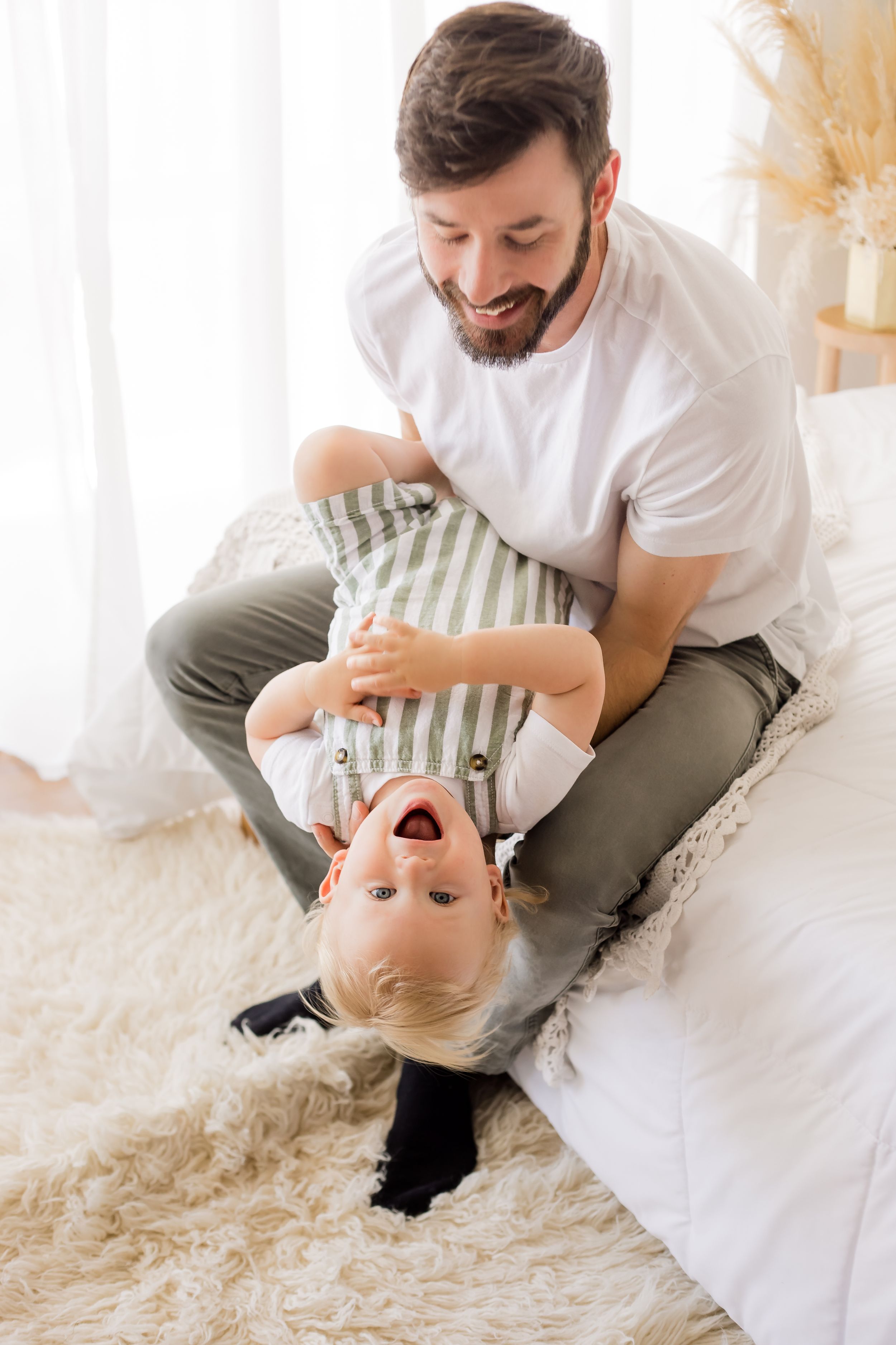 Dad playing with baby boy and laughing on the white bed in the white bedroom lifestyle room of the studio for their family portraits.