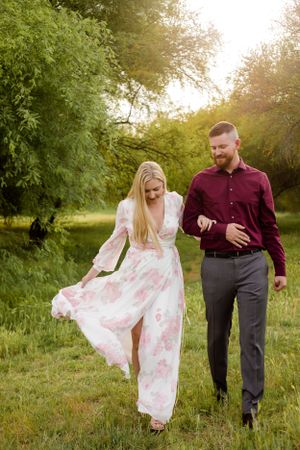Couple walking and laughing through a green field while her dress blows in the wind for their sweetheart outdoor Arizona session.