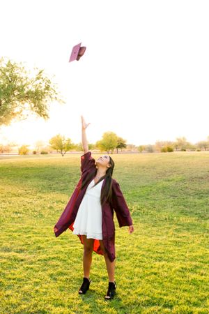 A girl throwing her cap in the air for a fun and creative photo during her cap and gown portrait graduate photoshoot on in a green field.