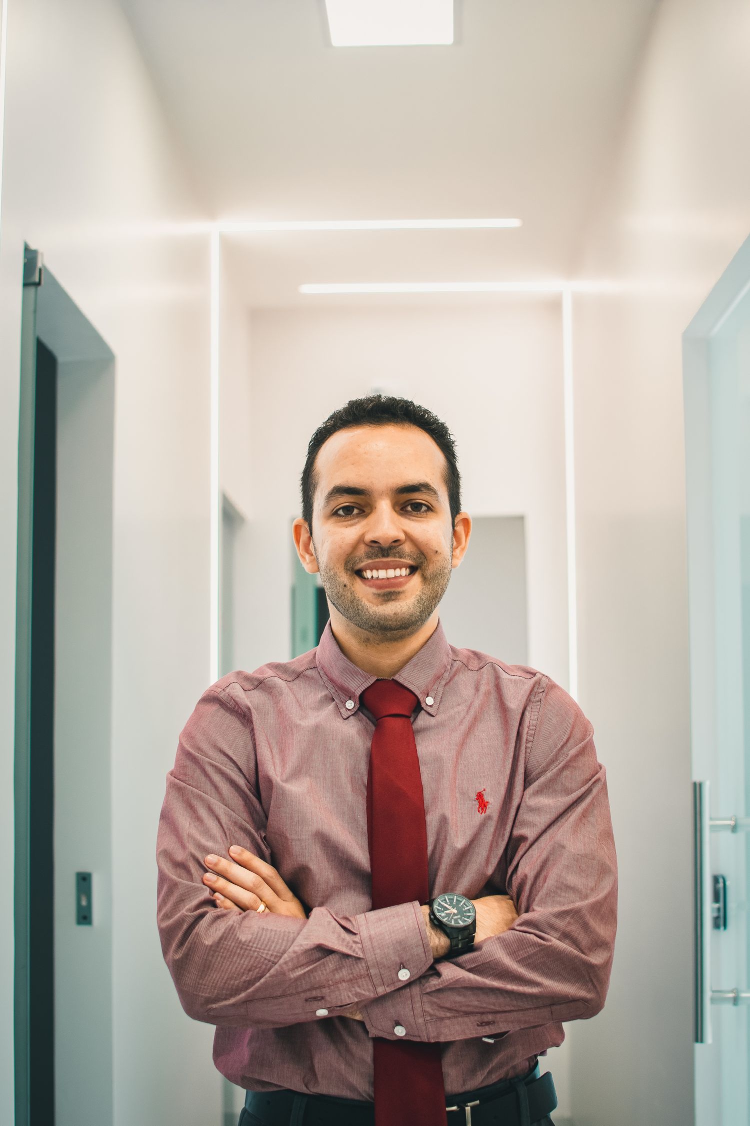 Medical professional in corridor wearing burgundy shirt and red tie with arms crossed.