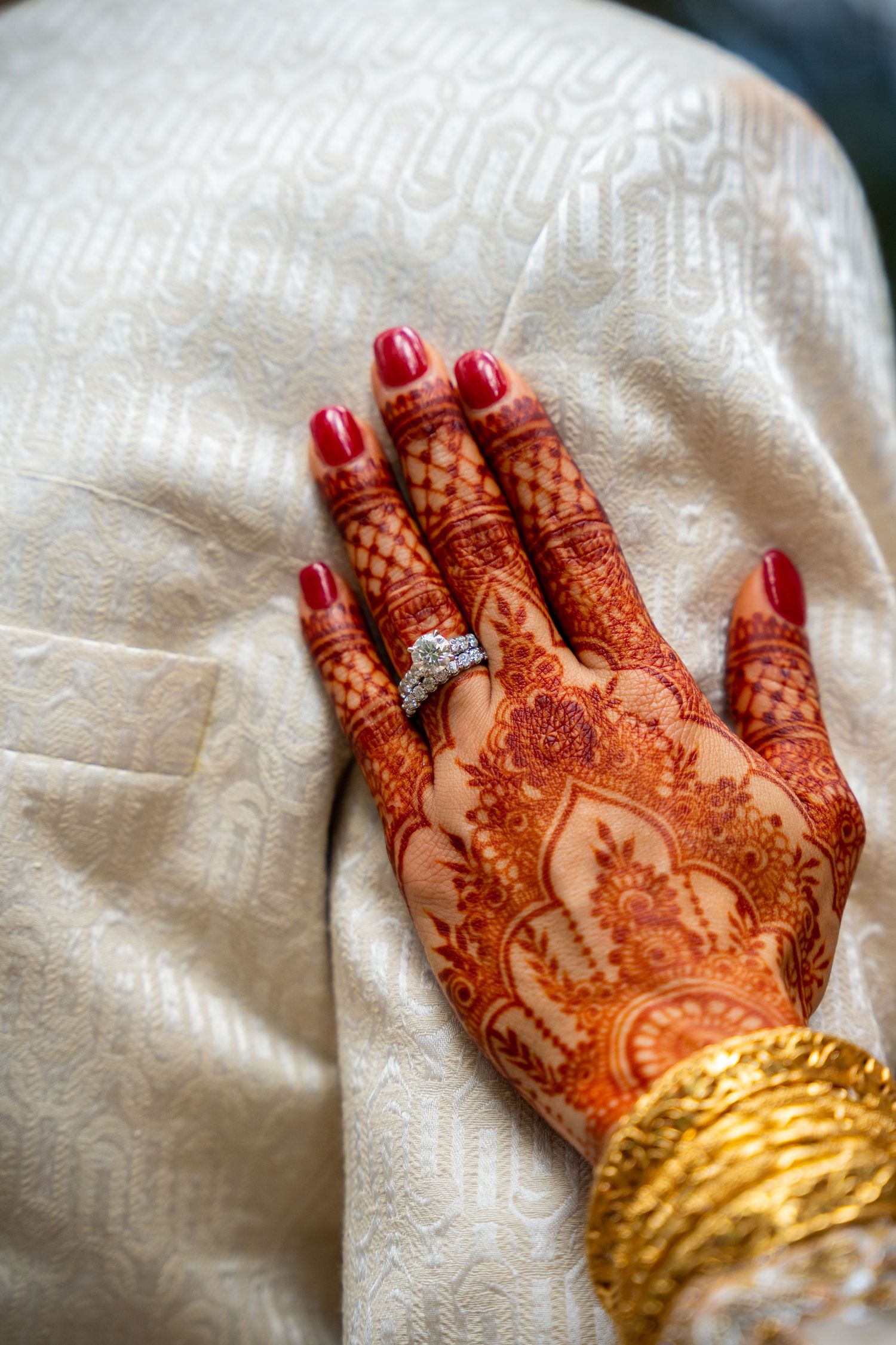 Detailed henna design on hand with red nails and engagement ring on white fabric.