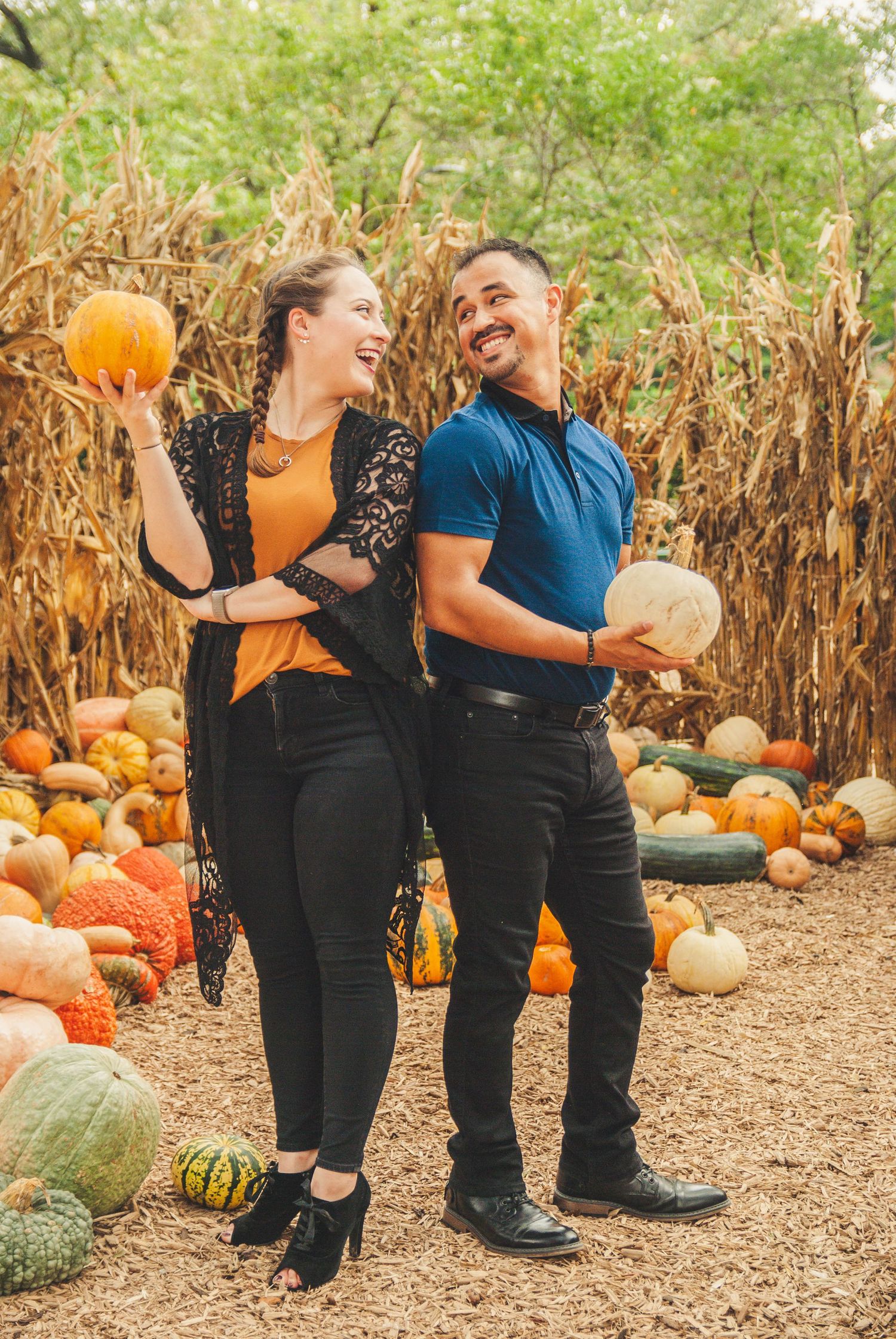 Smiling couple holding pumpkins in a festive fall pumpkin patch with corn stalks.