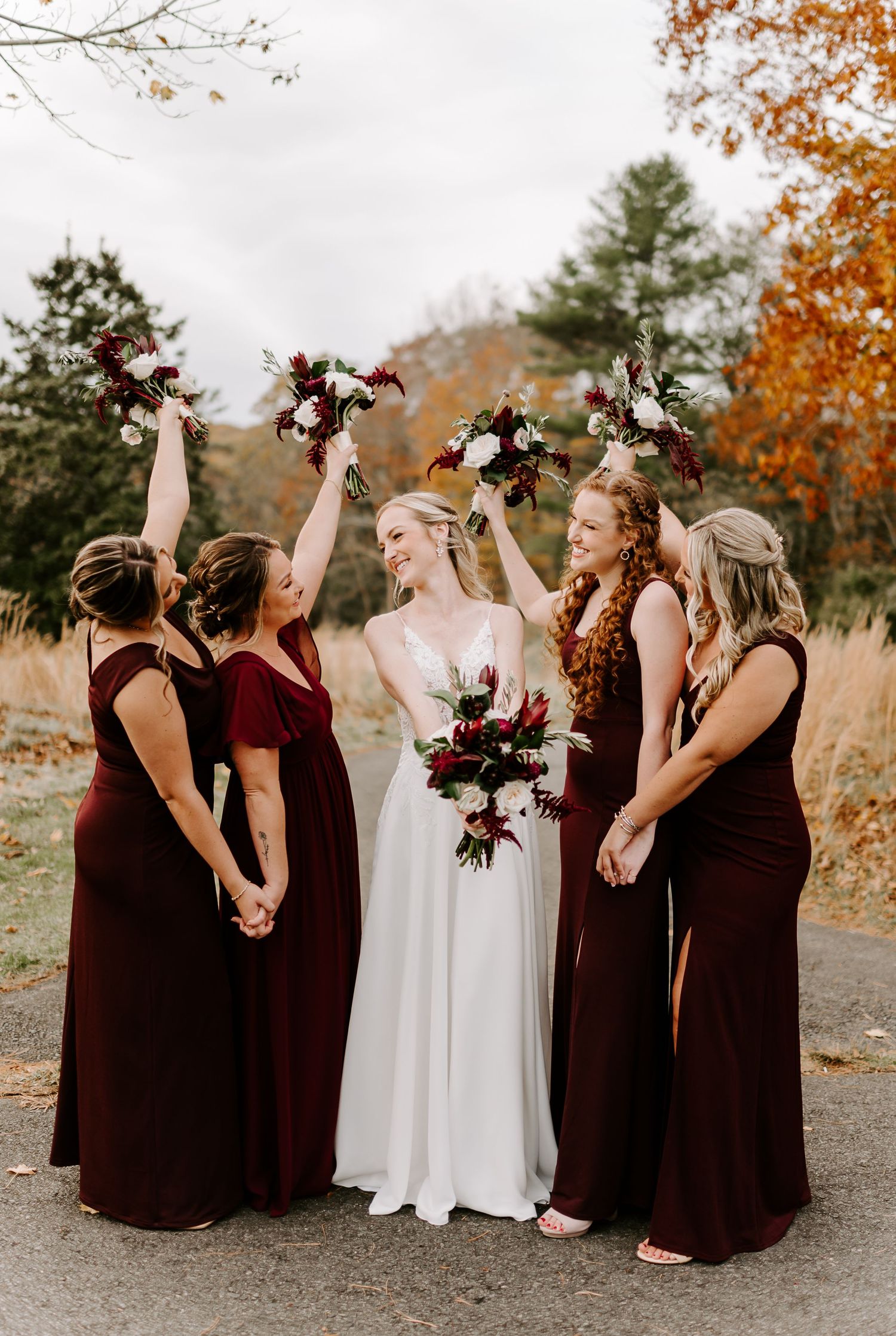 Bridesmaids in burgundy gowns raise bouquets with bride against fall foliage background.