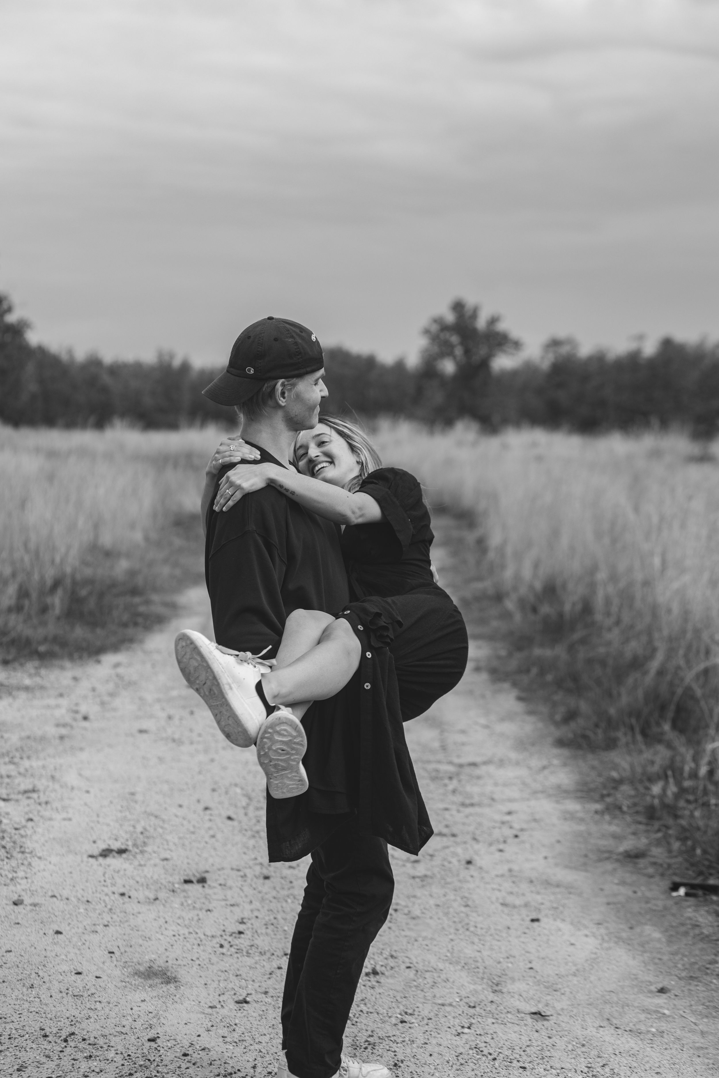 Black and white photo of person lifting partner in playful embrace on dirt path.