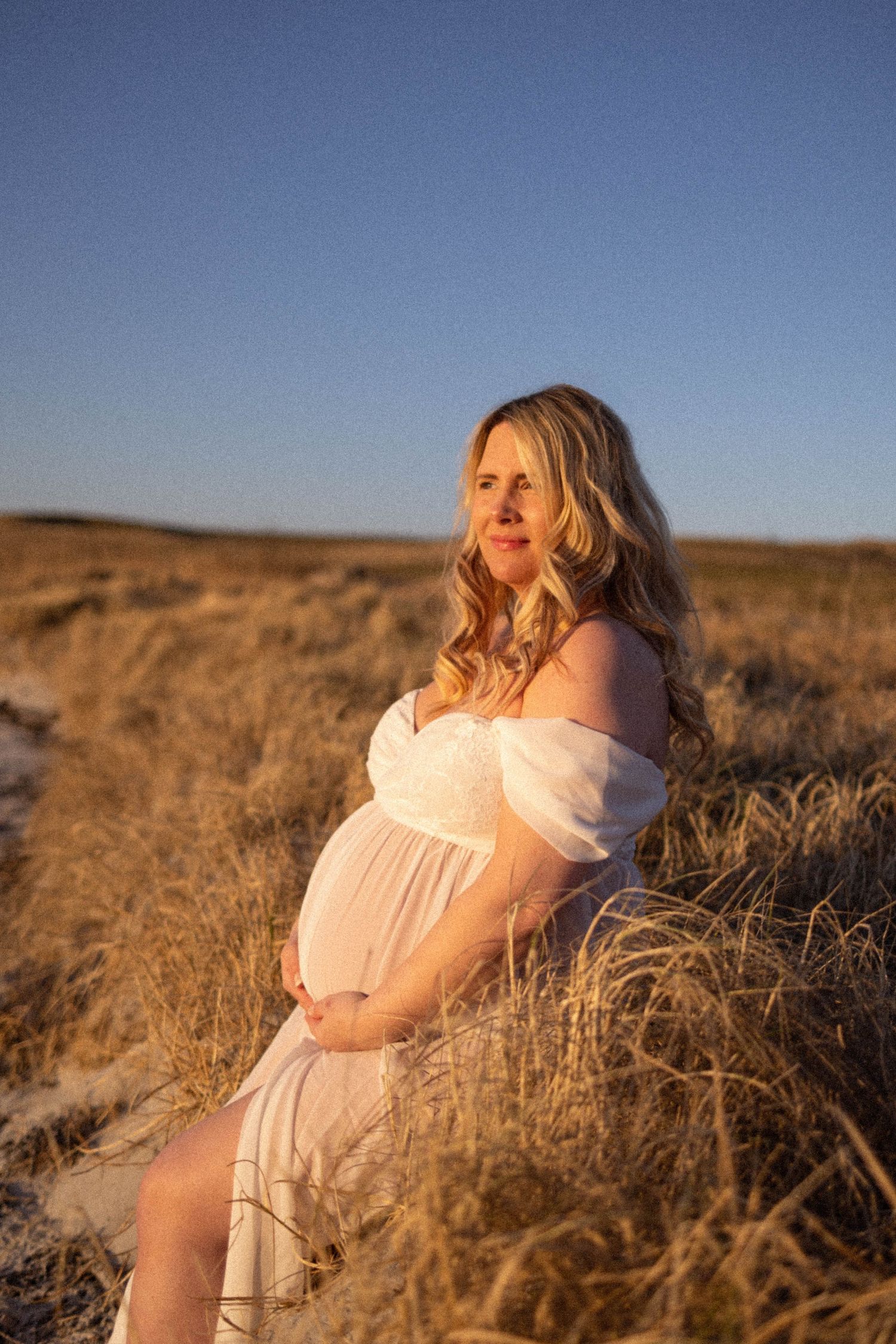Maternity photo of woman in white dress at sunset in golden field.