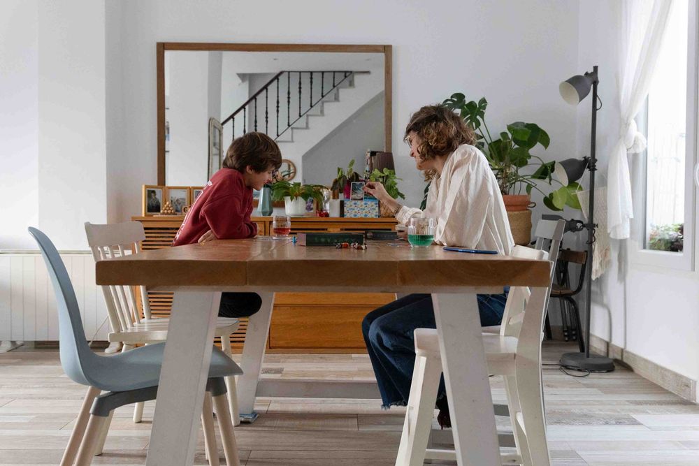 Two people chatting at modern white dining table with plants and staircase in background.