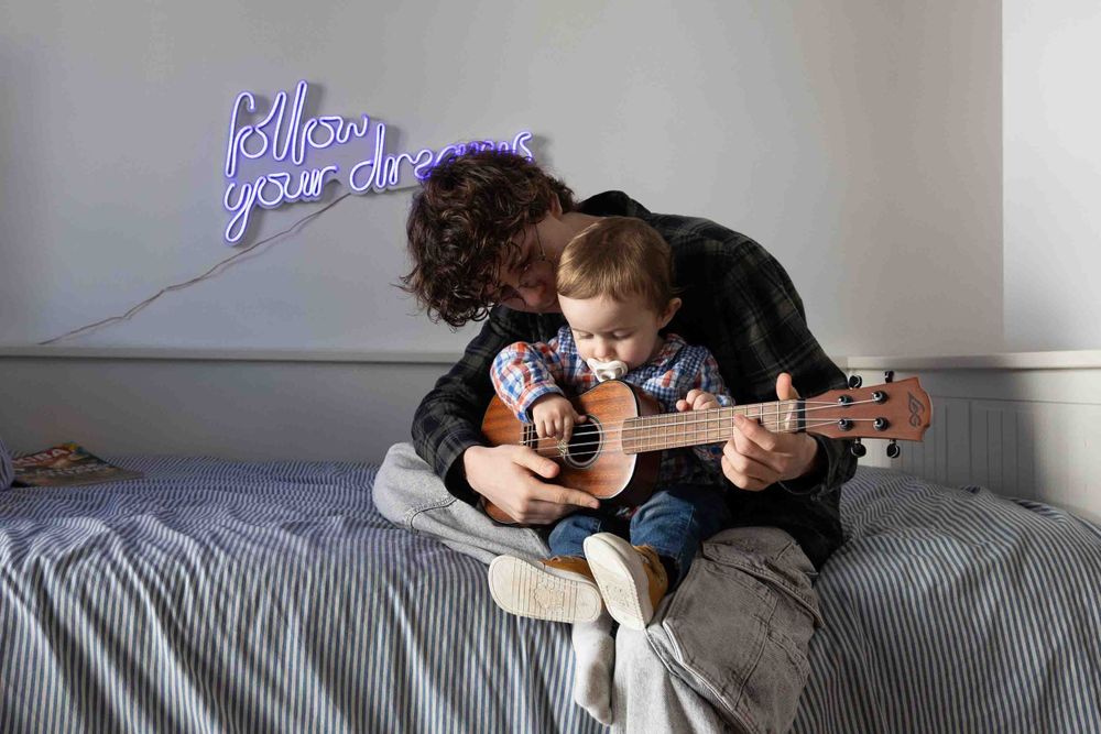 Adult and child playing ukulele together on a bed beneath a neon sign.