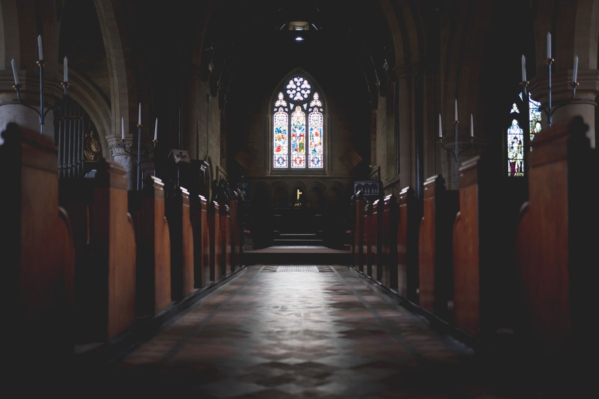 Dark church aisle lined with wooden pews leading to illuminated stained glass windows.
