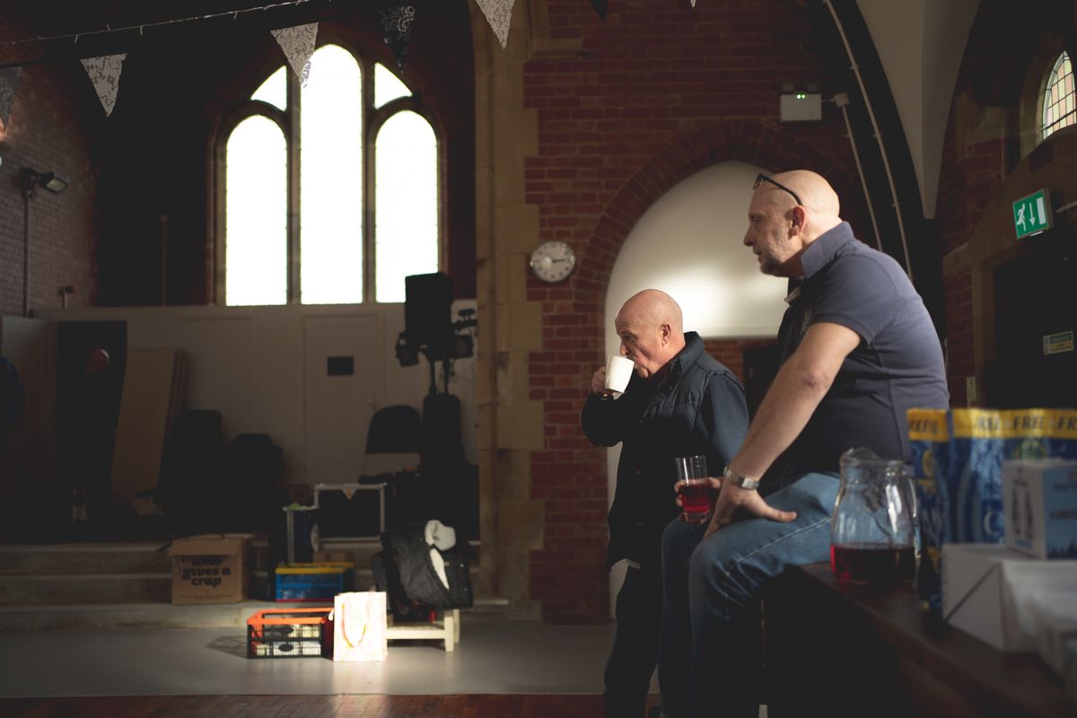 Two people sit in contemplation within a church or chapel with gothic arched windows and brick walls.