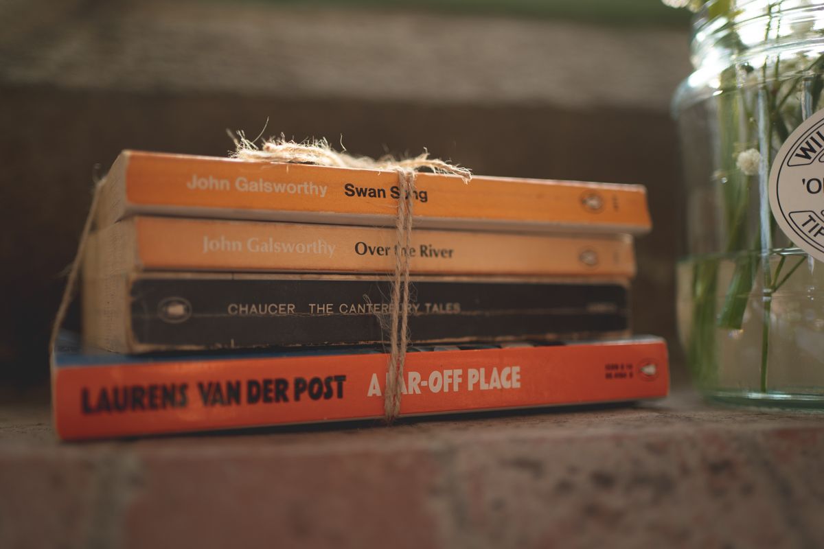 A stack of orange and red books lies on a wooden surface next to a glass jar.