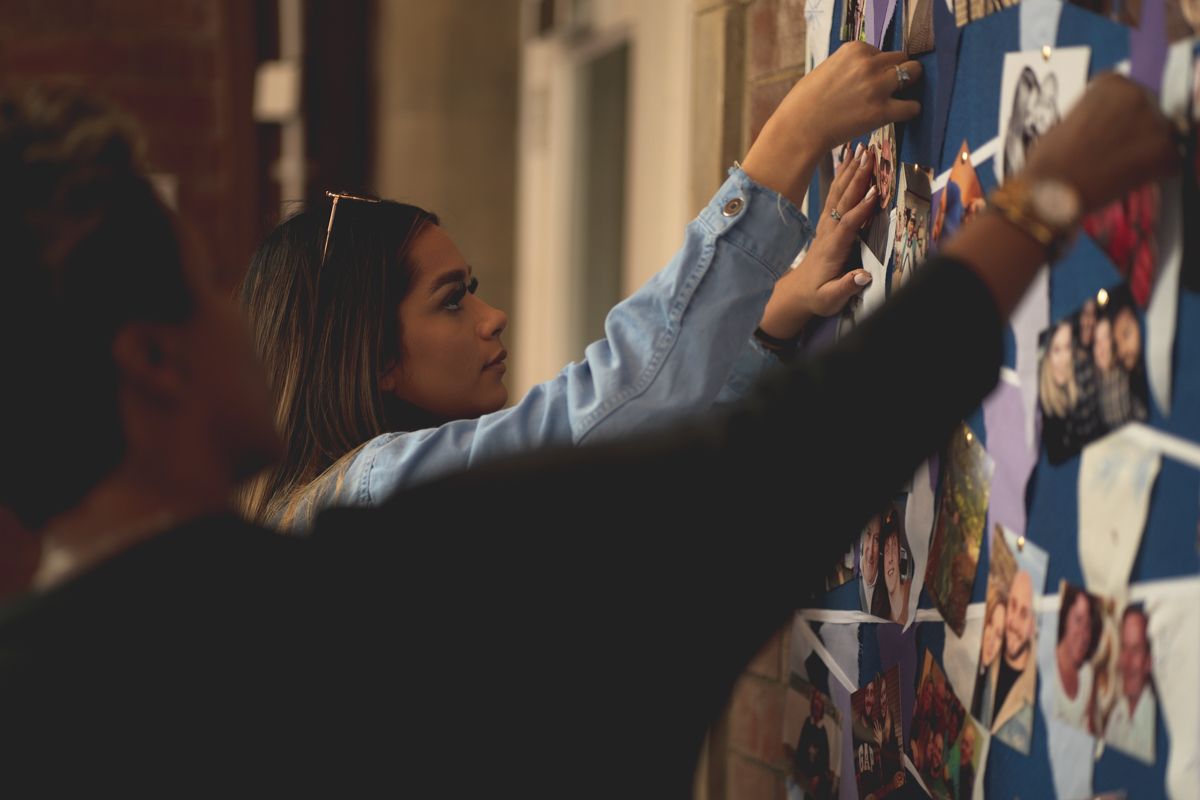 Someone reaches up to arrange photos on a display wall in dim lighting.