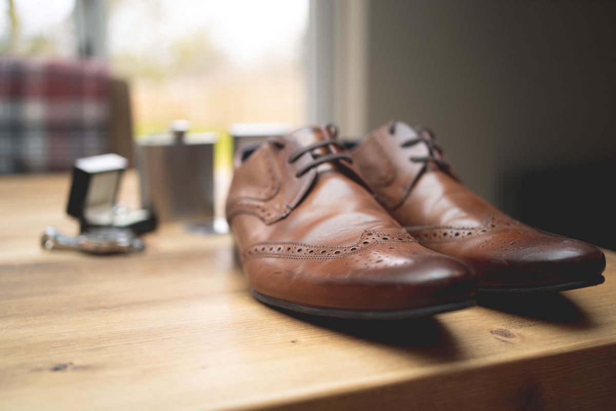 Brown leather wingtip dress shoes sit on a wooden surface next to a small object.