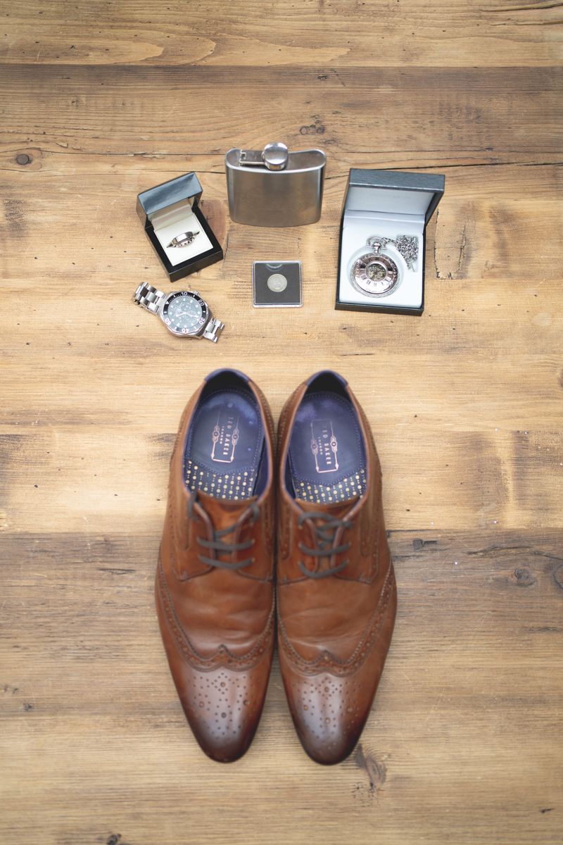 Wedding accessories including brown leather dress shoes, rings, watch and flask arranged on wooden surface.