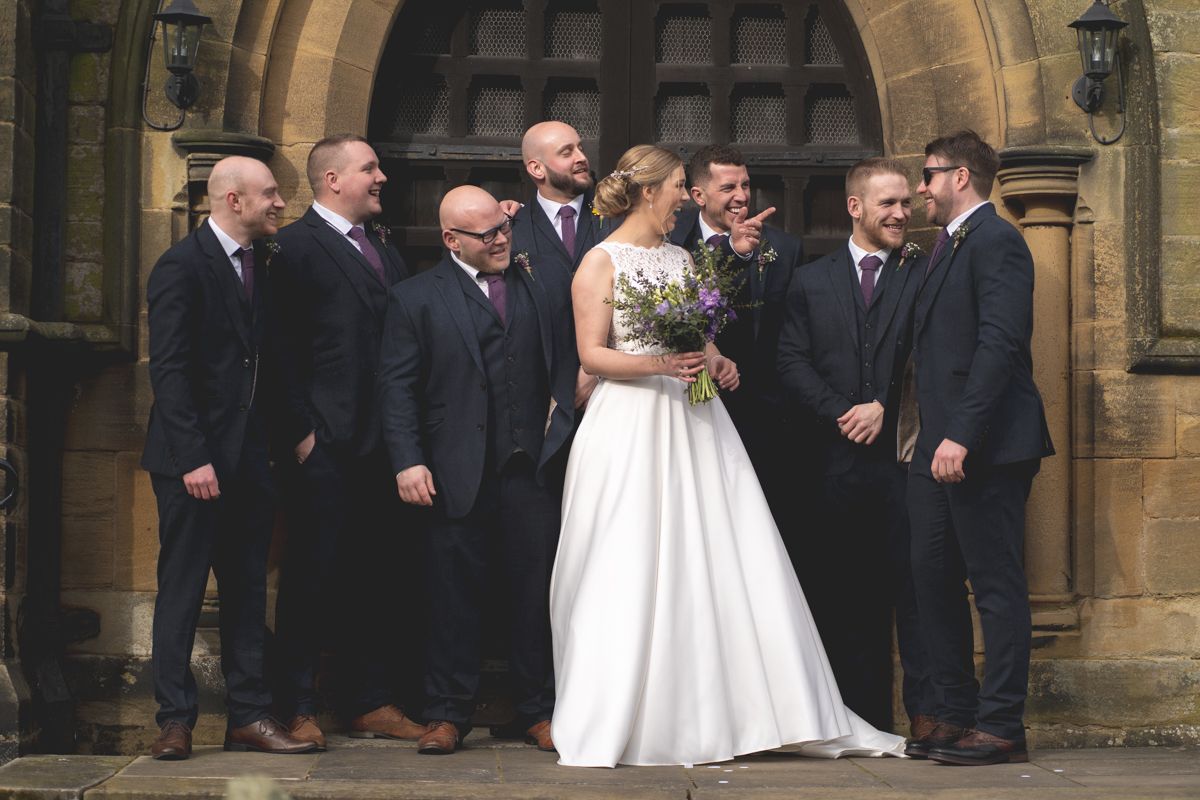 Wedding party members pose together against stone church architecture for a formal group photo.