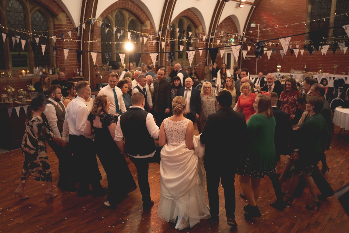 Wedding guests fill the dance floor of a historic venue decorated with string lights and bunting flags.