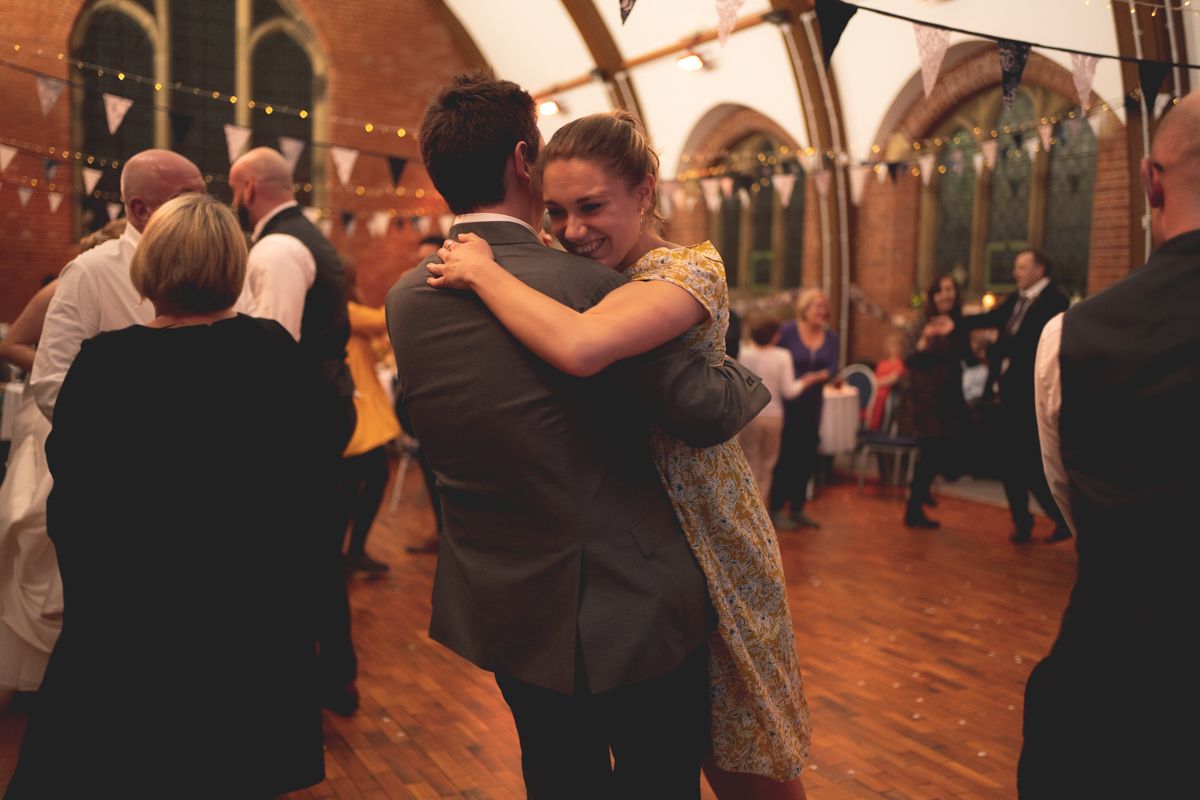 Couples dance and embrace on a wooden dance floor in a grand hall with arched windows.