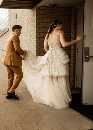 A couple enters a doorway with the bride's white flowing dress being held up by her partner in a brown suit.